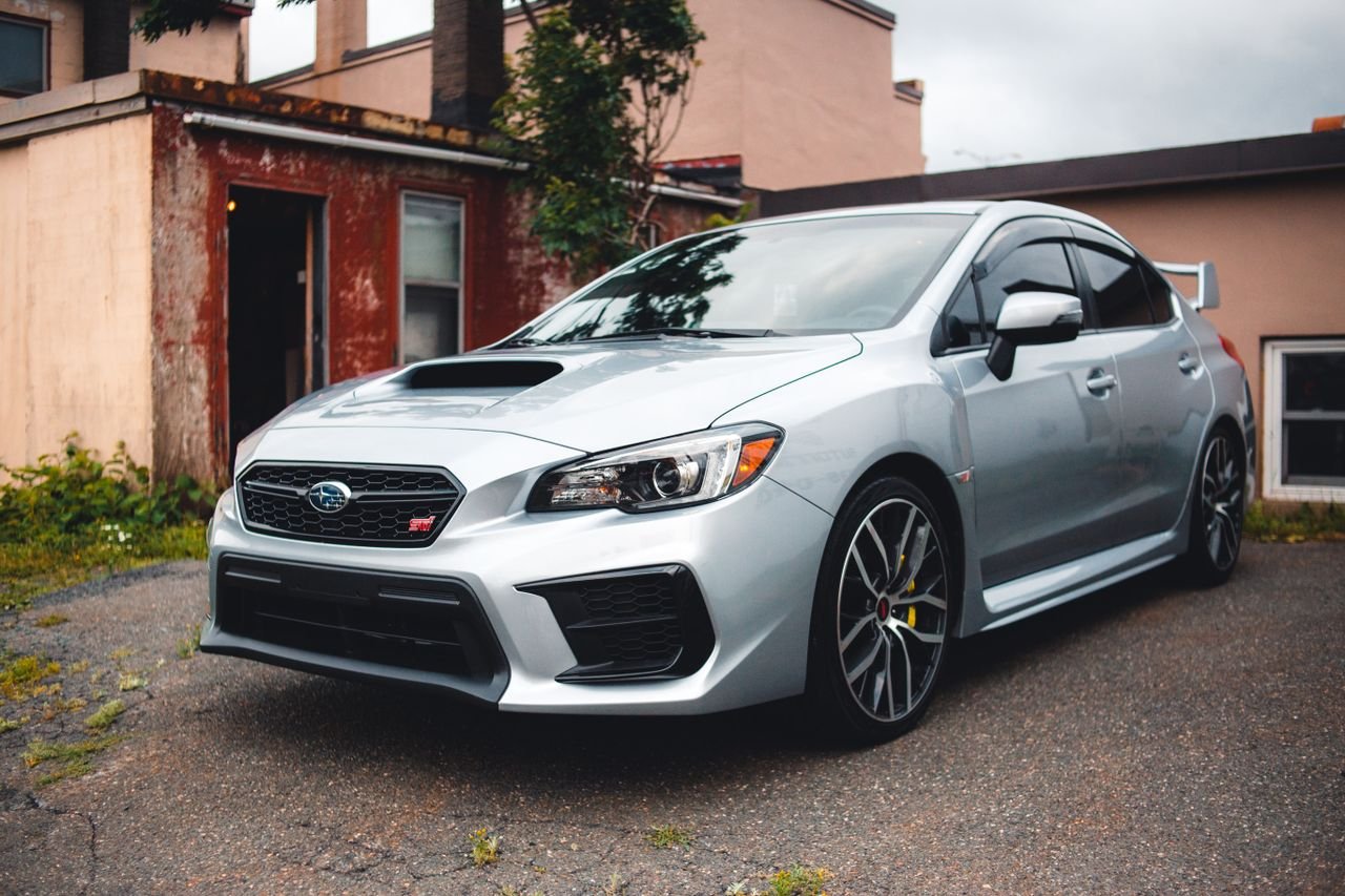 Silver Subaru WRX STI parked in a residential driveway with rustic backdrop.