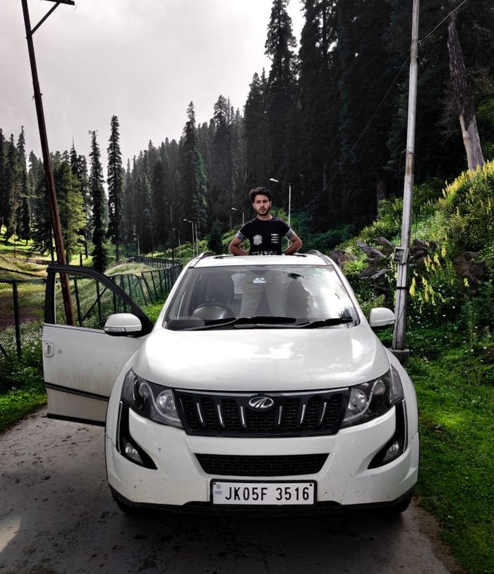 A white SUV parked outdoors with a person standing through the sunroof, surrounded by lush Kashmir scenery.